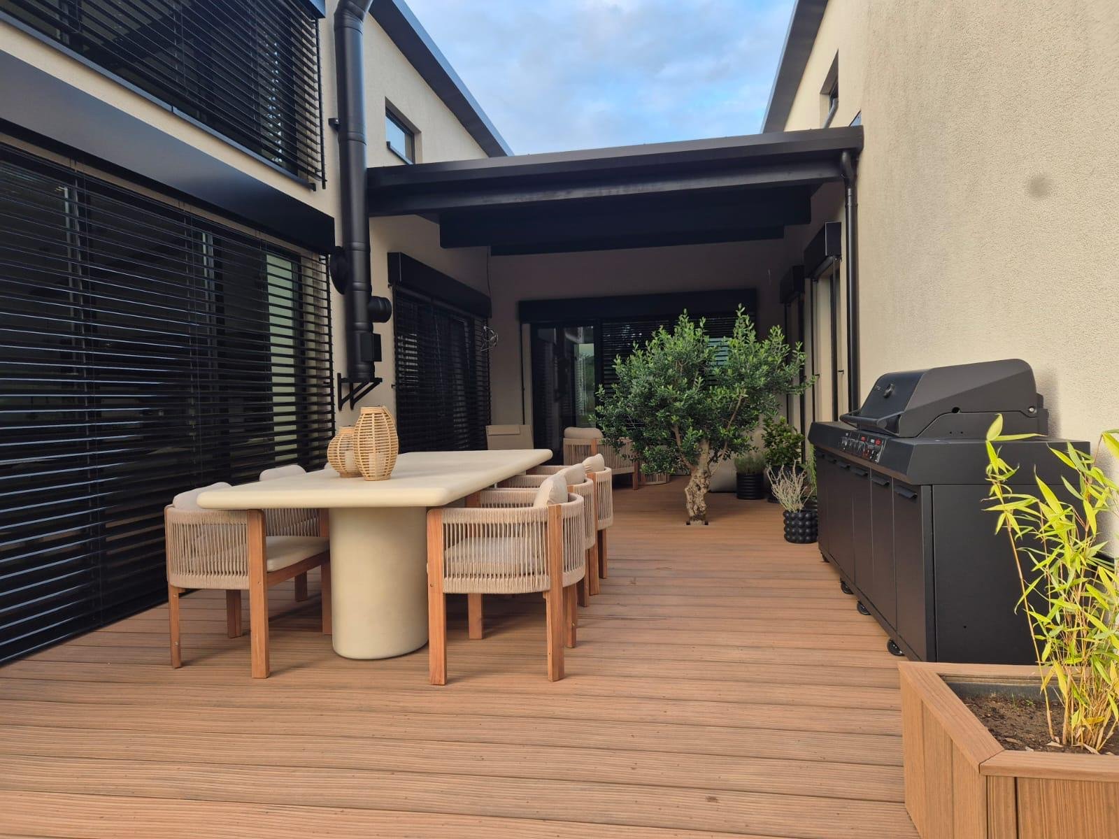 Überdachte Terrasse mit hellbraunen WPC-Dielen, langem Esstisch, sechs Korbstühlen, Olivenbaum und Gasgrill