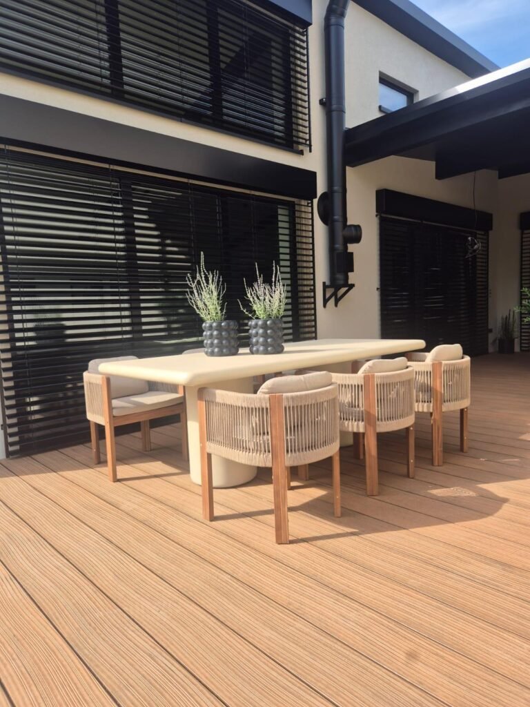 WPC-Terrasse in hellem Braunton bei Sonne, Esstisch mit sechs Stühlen vor einer Fassade mit schwarzen Raffstoren