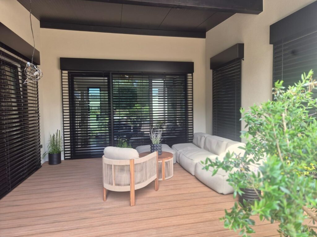 Überdachte Lounge auf hellbraunen WPC-Dielen mit hellem Sofa, Sessel und Pflanzen